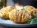 Drei goldbraune Hasselback Kartoffeln mit geschmolzenem Käse und Rosmarin auf einem blauen Teller.