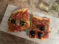 Zwei Scheiben Focaccia mit Tomaten, Knoblauch und schwarzen **Oliven Ligurien** garniert mit frischen Kräutern, daneben ein Glas Weißwein.
