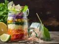 Bunter Salat im Glas mit Couscous, Karotten, Gurken, Paprika und Rotkohl. Daneben liegen Zitronen und Kräuter.