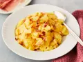 Ein Teller mit Kartoffelsalat mit Speck, garniert mit knusprigen Speckwürfeln. Daneben liegt ein Löffel.