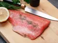 Frisches Lachsfilet mit Dill und Zitrone auf einem Holzbrett, bereit zum Kochen mit Gin. Ein scharfes Messer liegt daneben.