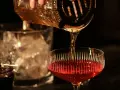 Ein **cocktail klassiker mit whisky und wermut** wird zubereitet. Bernstein-farbene Flüssigkeit fließt aus einem Shaker in ein Glas mit rotem Inhalt.