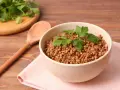 Eine Schüssel mit frisch gekochtem Buchweizen, garniert mit Petersilie. Daneben liegt ein Holzlöffel auf einer rosa Serviette.