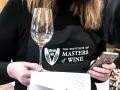 Eine Person mit einer Schürze des "Institute of Masters of Wine" hält ein Weinglas und ein Smartphone.
