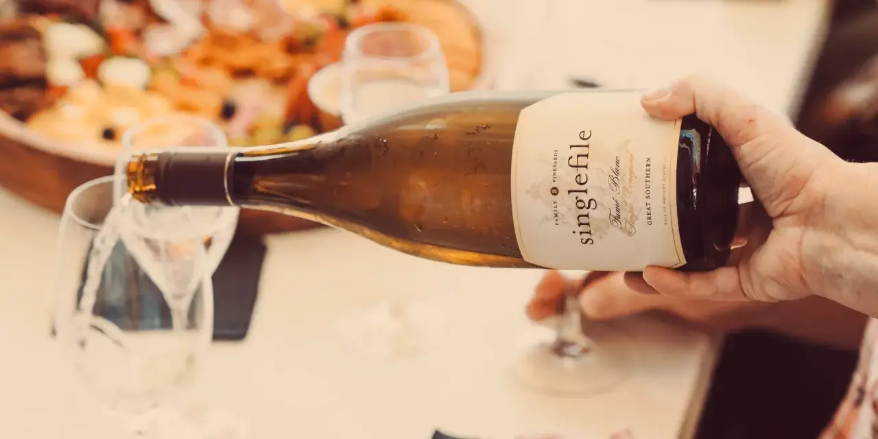 Eine Hand gießt einen **fumé wein** aus einer Flasche in ein Glas. Im Hintergrund ist eine Platte mit Speisen zu sehen.