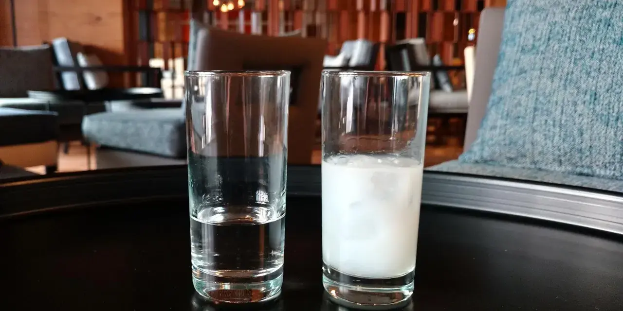 Zwei Gläser auf einem Tisch: eines mit klarem Wasser, das andere mit milchigem Ouzo. Ein Vergleich zwischen Raki und Ouzo.