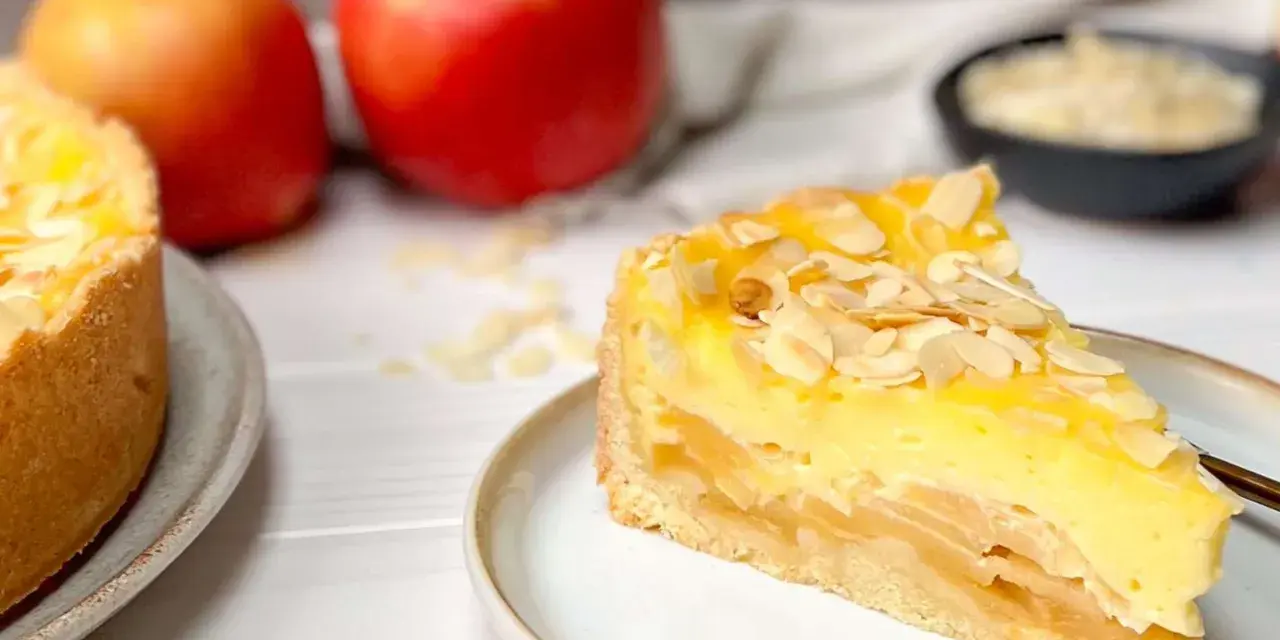Ein saftiges Stück Apfel Schmand Torte mit Mandelsplittern, daneben Äpfel und eine Schale mit Streuseln.