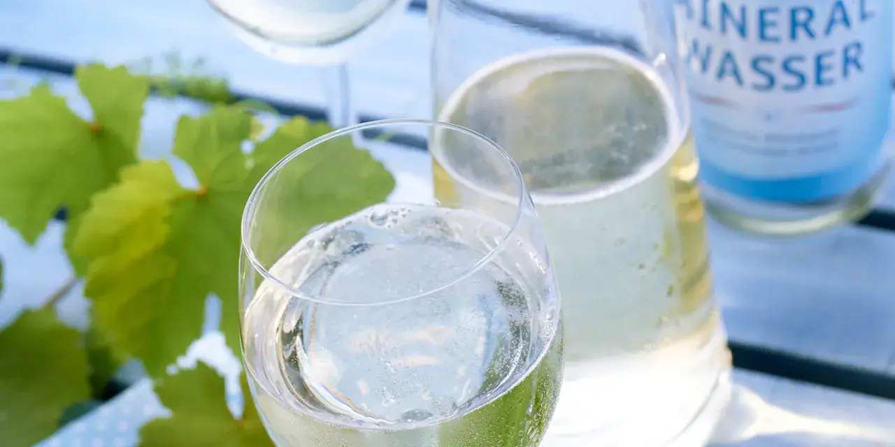 Erfrischendes Weinschorle-Rezept: Weißwein und Mineralwasser in Gläsern, daneben eine Karaffe und eine Flasche Mineralwasser. Grüne Weinblätter im Hintergrund.