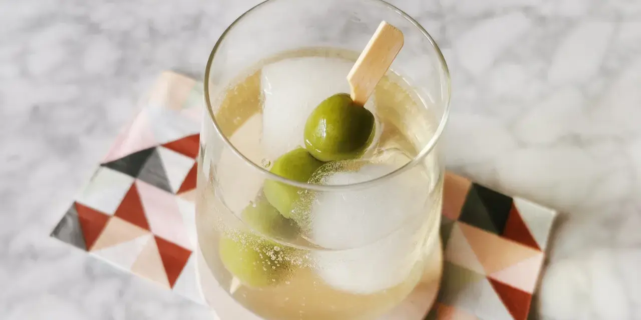 Erfrischender Italicus Spritz mit Eiswürfeln und Oliven auf einem gemusterten Untersetzer.