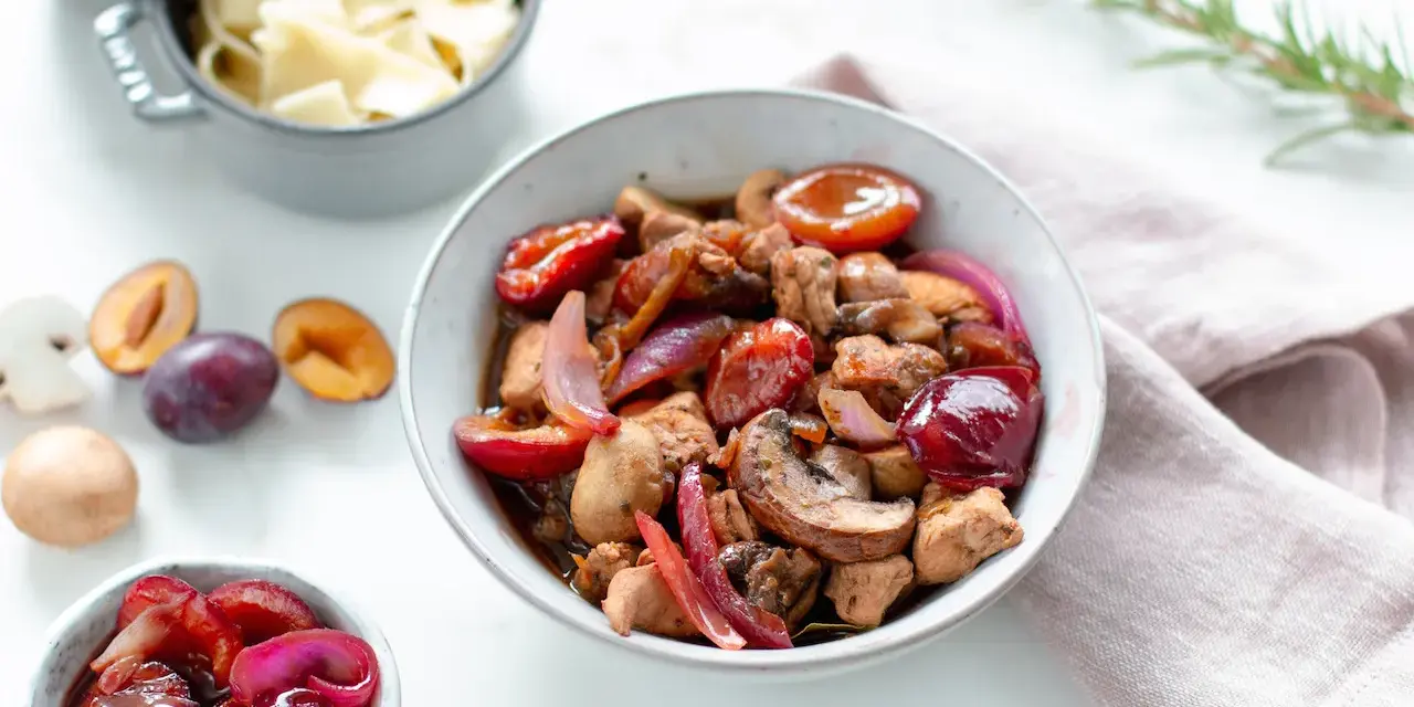Herzhafte Rezepte mit Pflaumen: Ein Eintopf mit Fleisch, Pilzen und roten Zwiebeln, garniert mit frischen Pflaumen. Daneben eine Schüssel mit Nudeln.