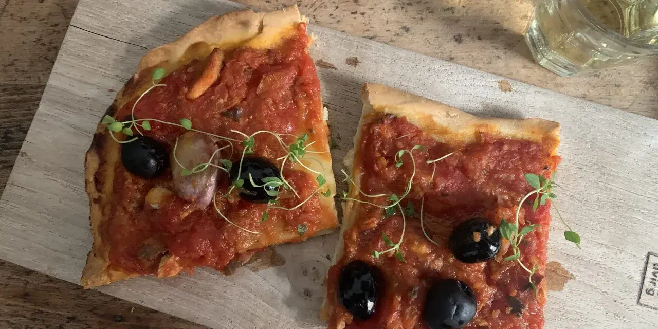 Zwei Scheiben Focaccia mit Tomaten, Knoblauch und schwarzen **Oliven Ligurien** garniert mit frischen Kräutern, daneben ein Glas Weißwein.