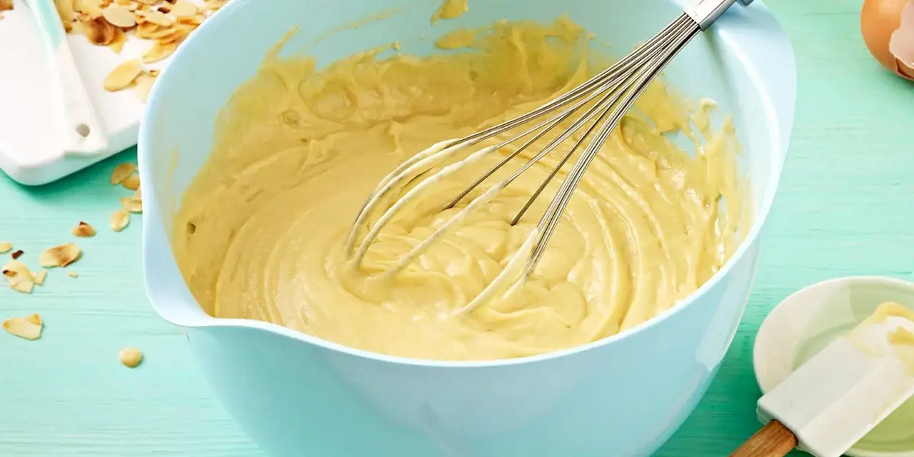 Ein hellblauer Schüssel mit Kuchenteig und Schneebesen. Daneben Mandelsplitter und Eier. Ein tolles Kuchenteig Rezept!