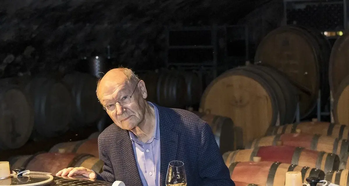 Ein Mann mit Brille hält ein Glas Wein in einem Weinkeller. Die Fässer im Hintergrund und der Wein lassen an Wein und Musik denken.
