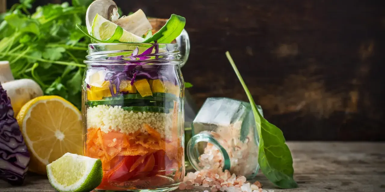 Bunter Salat im Glas mit Couscous, Karotten, Gurken, Paprika und Rotkohl. Daneben liegen Zitronen und Kräuter.