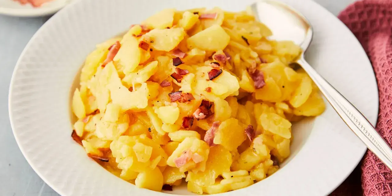 Ein Teller mit Kartoffelsalat mit Speck, garniert mit knusprigen Speckwürfeln. Daneben liegt ein Löffel.