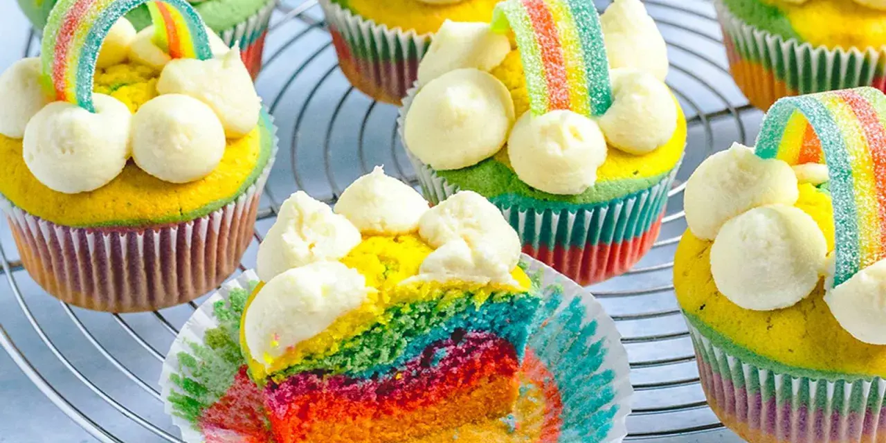 Bunte regenbogen muffins mit weißem Frosting und Gummibärchen-Regenbogen-Deko auf einem Gitter.