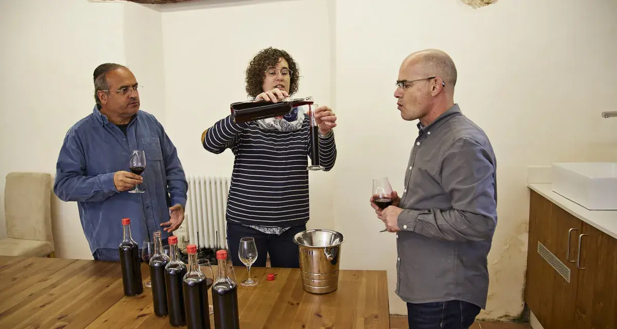 Weinprobe: Eine Frau schenkt Wein ein, während zwei Männer zuschauen. Auf dem Tisch stehen Flaschen für die Assemblage.