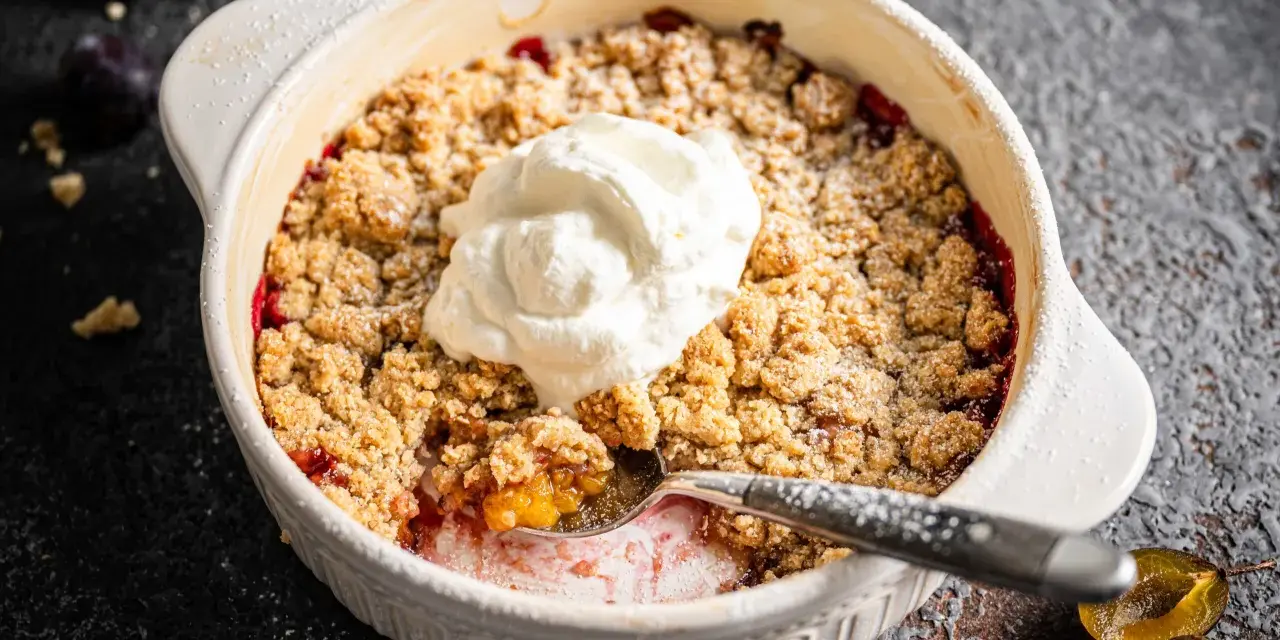 Ein warmer Zwetschgen Crumble mit Haferflocken, gekrönt von einem Klecks Sahne. Ein Löffel hebt die süße Frucht hervor.