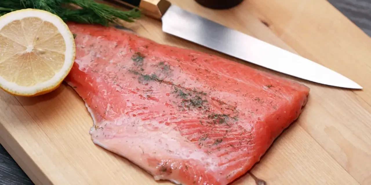 Frisches Lachsfilet mit Dill und Zitrone auf einem Holzbrett, bereit zum Kochen mit Gin. Ein scharfes Messer liegt daneben.