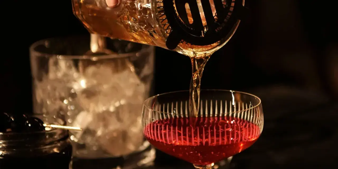 Ein **cocktail klassiker mit whisky und wermut** wird zubereitet. Bernstein-farbene Flüssigkeit fließt aus einem Shaker in ein Glas mit rotem Inhalt.
