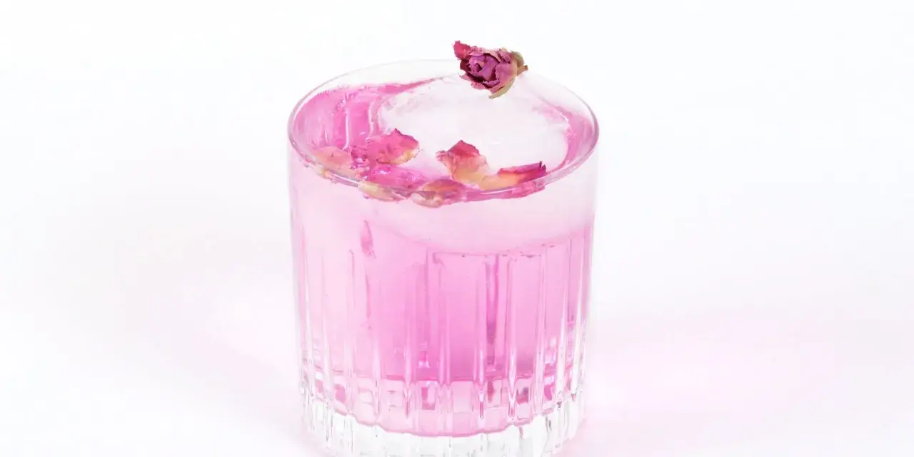 Ein erfrischender Gin Mix in zartem Rosa, garniert mit Rosenblättern und einer kleinen Knospe auf einem Eiswürfel.
