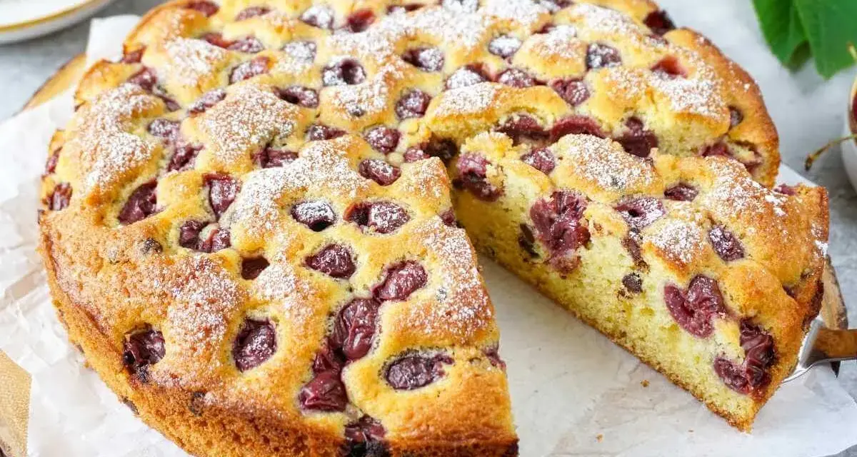 Ein saftiger, sommerlicher Kuchen mit Kirschen, bestäubt mit Puderzucker. Ein Stück wird gerade herausgeschnitten.