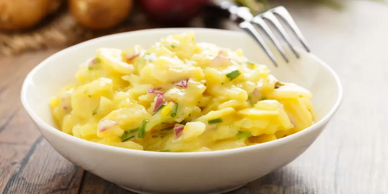 Ein cremiger Kartoffelsalat mit roten Zwiebeln und Schnittlauch in einer weißen Schüssel, bereit zum Genießen. Perfekt für ein Picknick oder als Beilage.