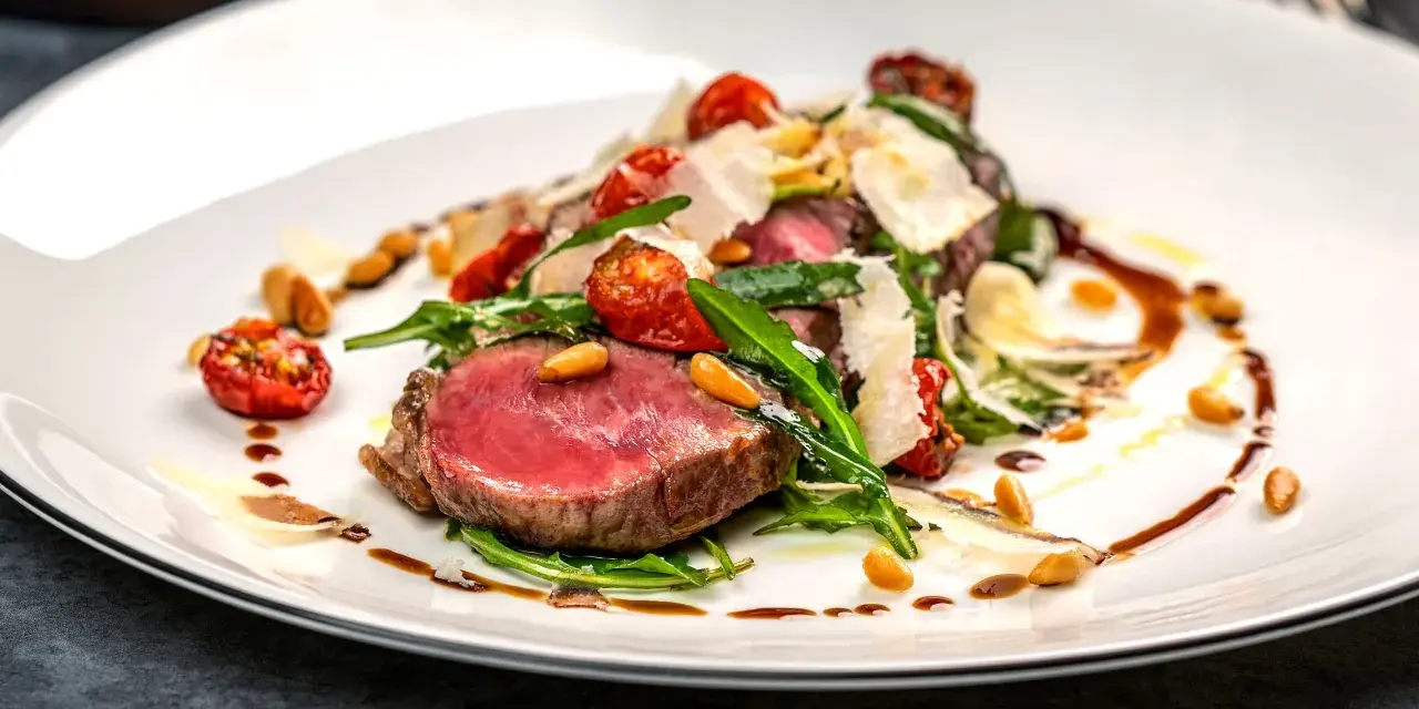 Saftiges Tagliata di Manzo mit Rucola, Kirschtomaten, Pinienkernen und Parmesan auf einem weißen Teller.