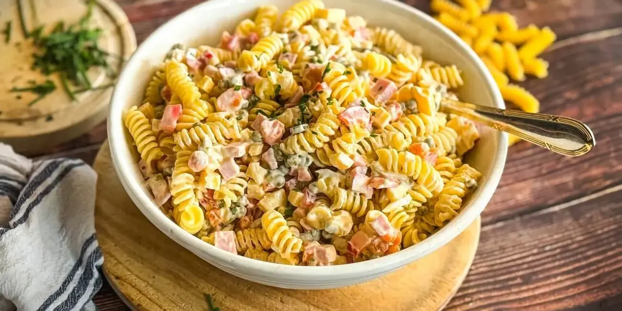 Ein cremiger Nudelsalat mit Schinken und Erbsen, ein Klassiker der klassischen Salate, serviert in einer weißen Schüssel auf einem Holztisch.