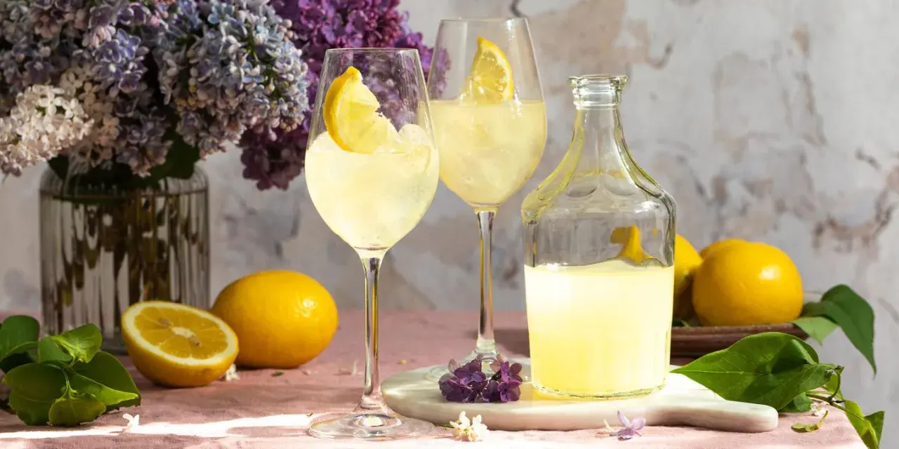 Zwei Gläser mit Eis und Zitronenscheiben, eine Flasche Limoncello und frische Zitronen auf einem rosafarbenen Tischtuch. Ein Hauch von Frühling mit Flieder.