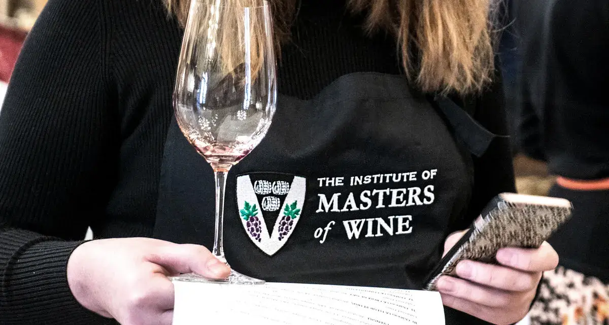 Eine Person mit einer Schürze des "Institute of Masters of Wine" hält ein Weinglas und ein Smartphone.
