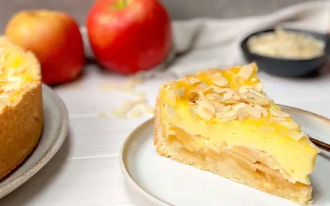 Ein saftiges Stück Apfel Schmand Torte mit Mandelsplittern, daneben Äpfel und eine Schale mit Streuseln.