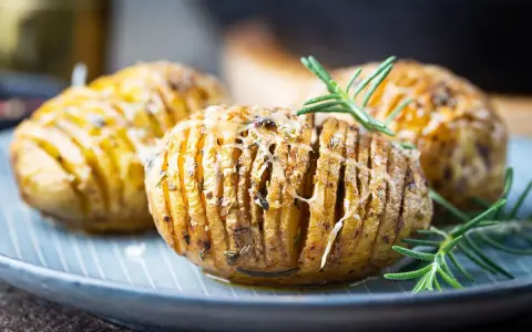 Drei goldbraune Hasselback Kartoffeln mit geschmolzenem Käse und Rosmarin auf einem blauen Teller.