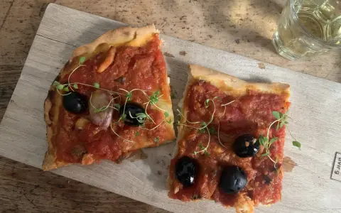 Zwei Scheiben Focaccia mit Tomaten, Knoblauch und schwarzen **Oliven Ligurien** garniert mit frischen Kräutern, daneben ein Glas Weißwein.