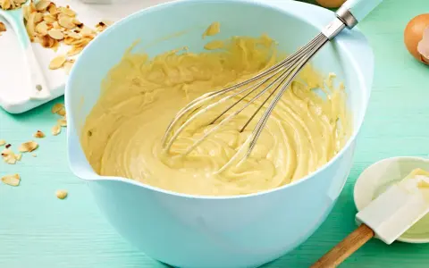 Ein hellblauer Schüssel mit Kuchenteig und Schneebesen. Daneben Mandelsplitter und Eier. Ein tolles Kuchenteig Rezept!
