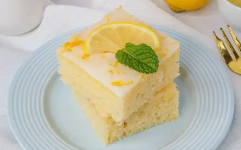 Zwei Stücke griechischer Zitronenkuchen mit Joghurt, garniert mit Zitronenscheibe und Minze, auf einem hellblauen Teller.