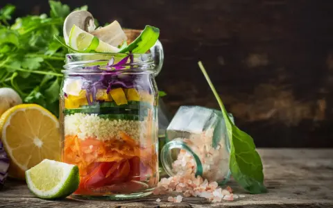 Bunter Salat im Glas mit Couscous, Karotten, Gurken, Paprika und Rotkohl. Daneben liegen Zitronen und Kräuter.