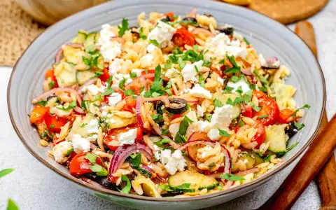 Bunter Kritharaki Salat mit Feta, Tomaten, Gurken, roten Zwiebeln und Oliven, garniert mit Petersilie. Ein köstlicher mediterraner Genuss.