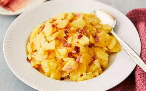 Ein Teller mit Kartoffelsalat mit Speck, garniert mit knusprigen Speckwürfeln. Daneben liegt ein Löffel.