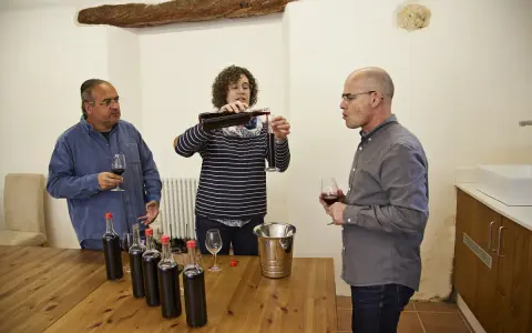Weinprobe: Eine Frau schenkt Wein ein, während zwei Männer zuschauen. Auf dem Tisch stehen Flaschen für die Assemblage.