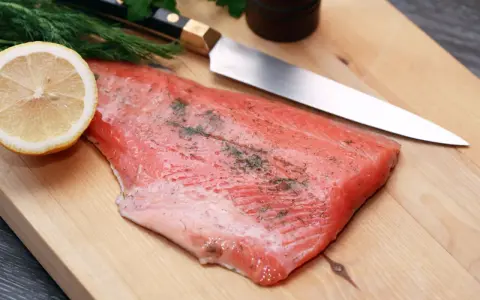 Frisches Lachsfilet mit Dill und Zitrone auf einem Holzbrett, bereit zum Kochen mit Gin. Ein scharfes Messer liegt daneben.