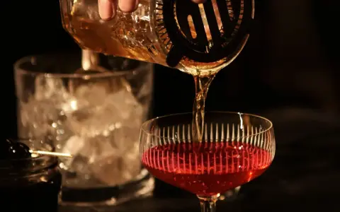 Ein **cocktail klassiker mit whisky und wermut** wird zubereitet. Bernstein-farbene Flüssigkeit fließt aus einem Shaker in ein Glas mit rotem Inhalt.