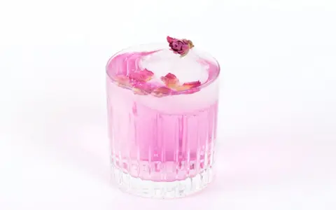 Ein erfrischender Gin Mix in zartem Rosa, garniert mit Rosenblättern und einer kleinen Knospe auf einem Eiswürfel.