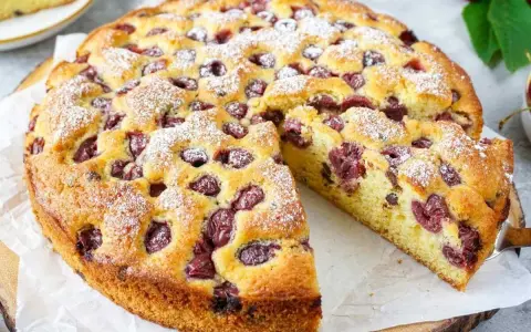 Ein saftiger, sommerlicher Kuchen mit Kirschen, bestäubt mit Puderzucker. Ein Stück wird gerade herausgeschnitten.