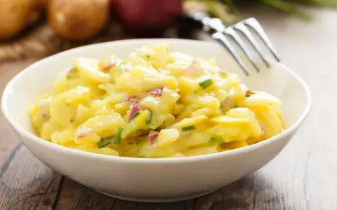 Ein cremiger Kartoffelsalat mit roten Zwiebeln und Schnittlauch in einer weißen Schüssel, bereit zum Genießen. Perfekt für ein Picknick oder als Beilage.
