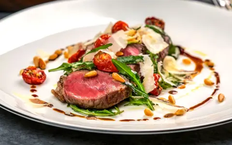 Saftiges Tagliata di Manzo mit Rucola, Kirschtomaten, Pinienkernen und Parmesan auf einem weißen Teller.