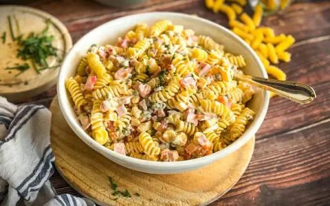 Ein cremiger Nudelsalat mit Schinken und Erbsen, ein Klassiker der klassischen Salate, serviert in einer weißen Schüssel auf einem Holztisch.
