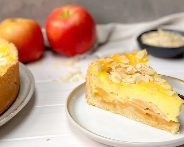 Ein saftiges Stück Apfel Schmand Torte mit Mandelsplittern, daneben Äpfel und eine Schale mit Streuseln.