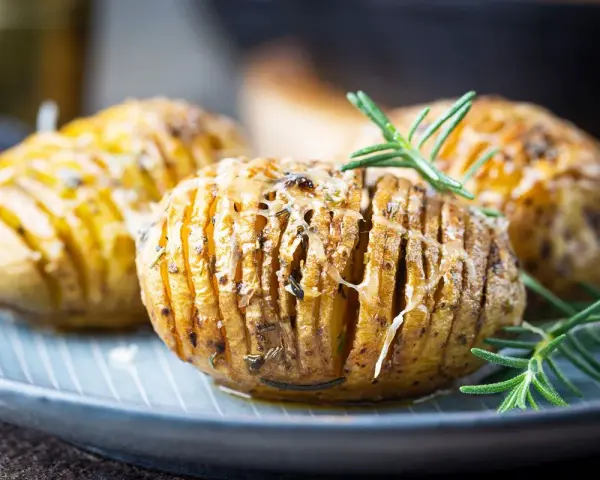 Drei goldbraune Hasselback Kartoffeln mit geschmolzenem Käse und Rosmarin auf einem blauen Teller.
