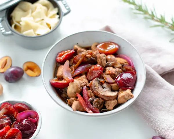 Herzhafte Rezepte mit Pflaumen: Ein Eintopf mit Fleisch, Pilzen und roten Zwiebeln, garniert mit frischen Pflaumen. Daneben eine Schüssel mit Nudeln.